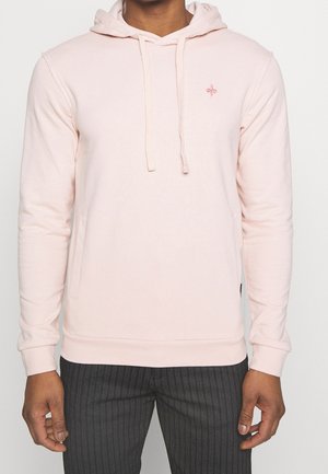 Mann trägt hellrosa Kapuzenpullover mit kleinem Logo auf der Brust und dunkelgraue Nadelstreifenhose, steht mit entspannten Armen am Körper.