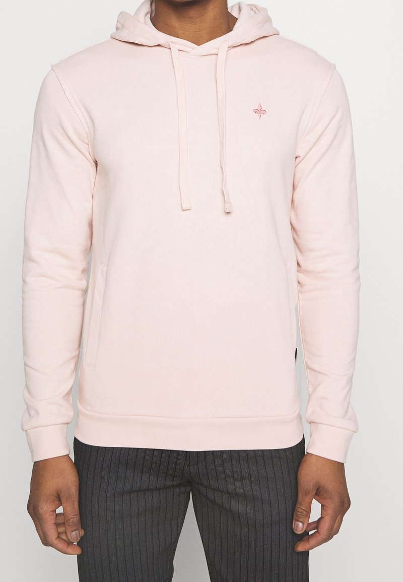 Homme portant un sweat à capuche rose clair avec un petit logo sur la poitrine et un pantalon gris foncé à fines rayures, debout, bras détendus le long du corps.