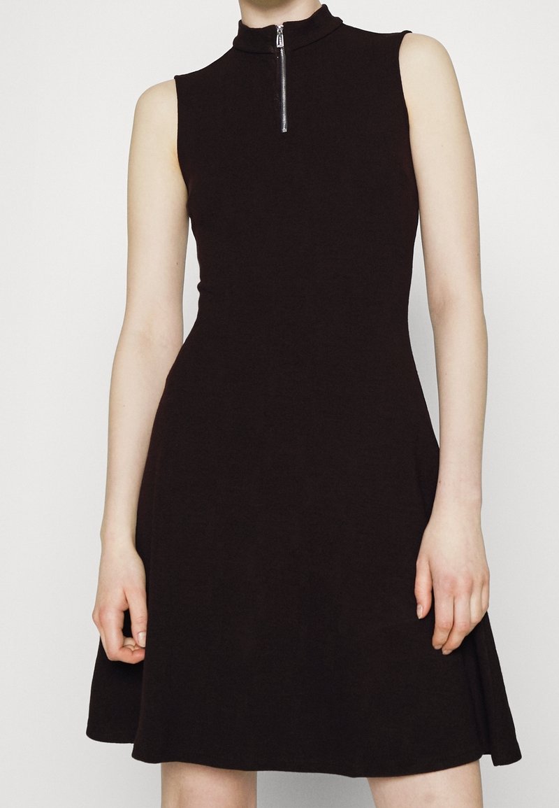 Robe noire sans manches avec col montant et fermeture éclair à l'avant, cintrée à la taille avec une jupe légèrement évasée, portée par une personne debout.