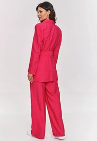Jasnoróżowy garnitur z dopasowaną marynarką z paskiem i szerokimi spodniami. Gładka tekstura, ostre wykończenie kołnierza oraz casualowe białe sneakersy.