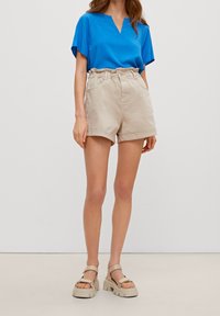 Kobieta ubrana w niebieską bluzkę z dekoltem w serek i krótkim rękawem, beżowe szorty z wysokim stanem i podwiniętym wykończeniem, oraz beżowe sandały na grubej podeszwie.