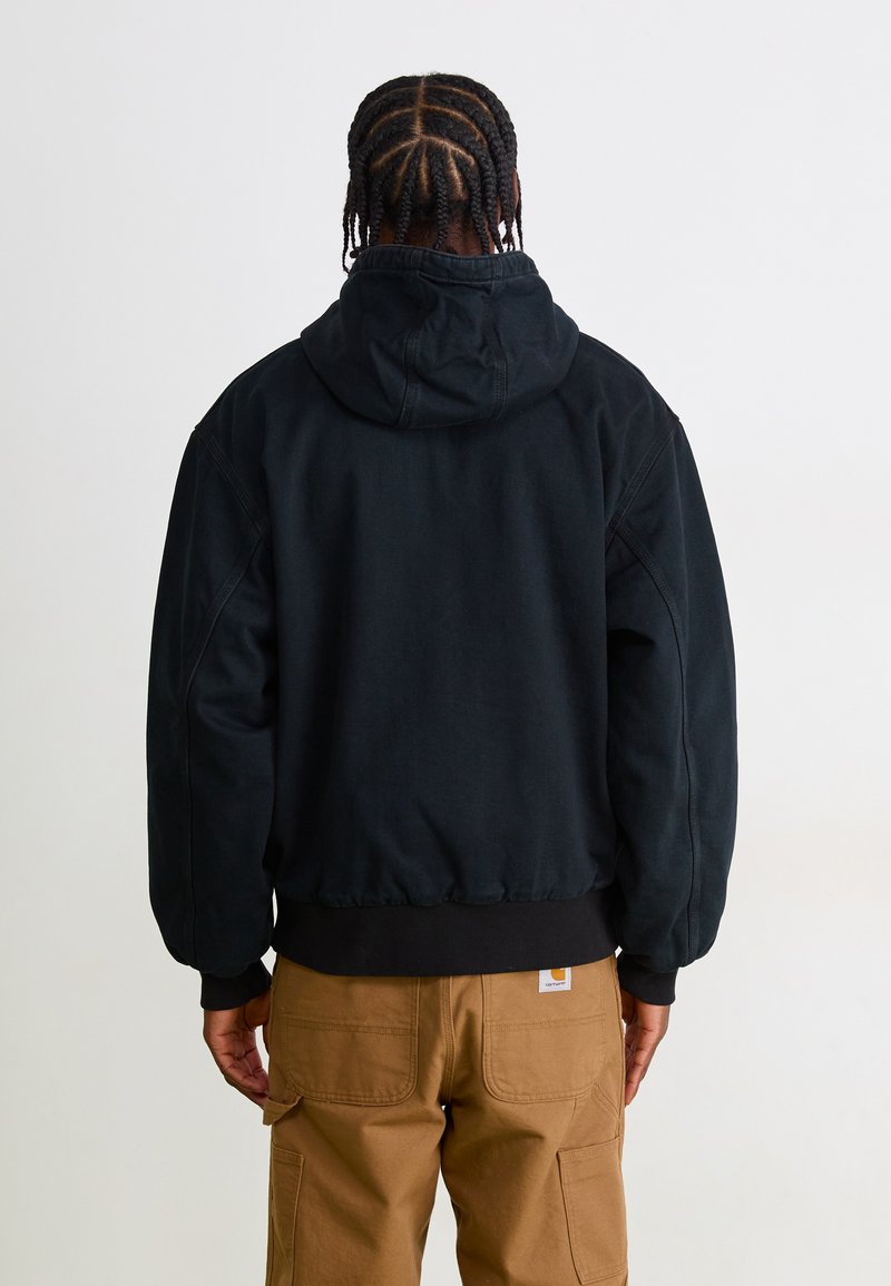 Person mit geflochtenen Haaren, die eine schwarze Kapuzenjacke und braune Cargo-Hosen trägt, steht mit dem Rücken zu einem einfarbigen Hintergrund.