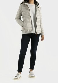 Lysegrå polstret jakke med hætte, frontlynlås, to sidelommer, båret over en grå sweater og mørke jeans, kombineret med hvide sneakers.