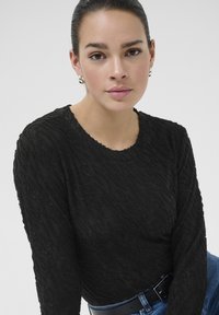 Sort langærmet top med struktureret bølget mønster, rund halsudskæring og let frynsede kanter, parret med blå denim og et sort bælte.