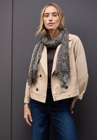 Giacca beige doppiopetto con bottoni neri, abbinata a un top nero e a una sciarpa grigia con motivo leopardato nero, su uno sfondo grigio.