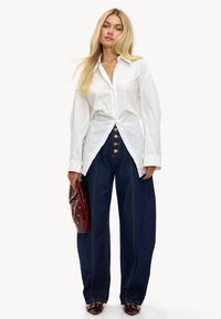 Camisa blanca abotonada con cuello puntiagudo, combinada con jeans de mezclilla oscura de pierna ancha, que presentan grandes botones delanteros y una textura suave.