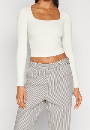 Vrouw draagt een witte, cropped top met lange mouwen en een vierkante halslijn, gecombineerd met een hoge taille broek met zwart-witte verticale strepen.