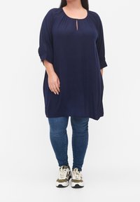 Marinblå tunika med rund halsringning och nyckelhål. Har trekvartsärmar och sidofickor, i kombination med jeans och sneakers.