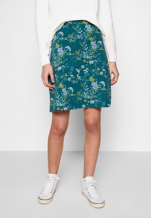 Jupe bleu sarcelle longueur genou avec motifs floraux et d'oiseaux, portée avec des baskets blanches montantes et un haut blanc à manches longues.
