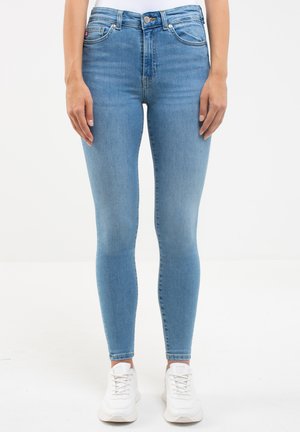Hellblaue Skinny-Jeans mit hoher Taille, aus Denim, mit fünf Taschen und einem Reißverschluss-/Knopfverschluss, kombiniert mit weißen Sneakers.