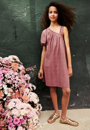 Niña joven con cabello rizado, vestida con un vestido malva de un solo hombro, sosteniendo una flor y de pie junto a un gran ramo de flores rosas y blancas.