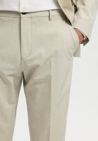 Pantalon de costume beige porté avec une veste assortie et une chemise blanche, main placée dans la poche droite, gros plan sur le bas du torse.