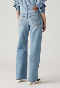 Light blue denim jeans with a high waist, straight leg, visible back pockets, and a brown leather patch featuring logo at the back waistband.