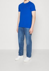 Blå kortärmad t-shirt i bomull med rund hals och en liten logotyp på bröstet, ihop med ljusblå jeans och vita sneakers.