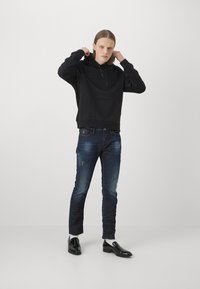 Fekete kapucnis pulóver, dombornyomott logóval, vékony szabású sötétkék farmernadrággal, amelyen enyhe kopás és elhasználtság látható. Fekete cipő.