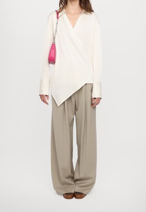 Vrouw die een crèmekleurige asymmetrische blouse draagt, wijde beige broek, bruine schoenen en een kleine roze schoudertas met kettingriem meeneemt.