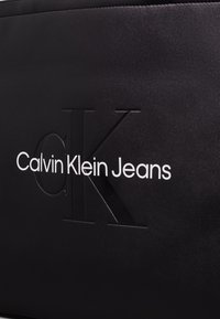 Czarna teksturowana powierzchnia z wytłoczonym logo "CK" oraz napisem "Calvin Klein Jeans" w białej czcionce bezszeryfowej, tworząca elegancki kontrast.
