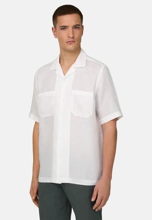 Junger Mann mit kurzen Haaren, der ein weißes, kurzärmeliges Hemd mit Knöpfen und zwei Brusttaschen sowie dunkle Hosen vor einem schlichten Hintergrund trägt.