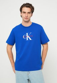 Modré bavlnené tričko s bielym a červeným logom "CK" na prednej strane. Klasický okrúhly výstrih a krátke rukávy, v kombinácii so svetlomodrými džínsami.