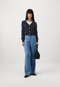 Námořnický svetr s kabelkovým pletením a pěti stříbrnými knoflíky, spárovaný se světle modrými širokými džínami a texturovanou černou kabelkou.