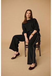 Vrouw zittend op een houten stoel met een zwarte broek met wijde pijpen en witte bies, bijpassende losse zwarte top, bruine armband en donkerbruine platte schoenen.