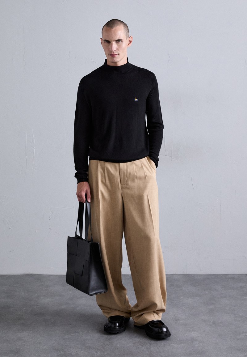 Mann trägt schwarzen Langarmpullover, beigen weiten Hosen, schwarze Schuhe, hält eine schwarze Leder-Tote-Bag und steht vor einer schlichten grauen Wand.