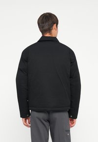 Black, padded jacket with a classic collar, button cuffs, and a boxy fit, displayed from the back. Textured finish with a minimal design.