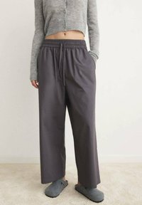 Persoon in losse donkergrijze broek met elastische tailleband en trekkoord, grijze cropped cardigan en grijze instapslippers, staande binnenshuis.