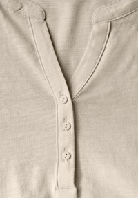 Hellbeige, weiches Stoffhemd mit V-Ausschnitt, drei weißen Knöpfen und einem runden Kragen mit zarten Stickdetails.