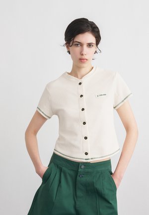 Femme portant un haut blanc à manches courtes boutonné avec des boutons noirs et un short vert, debout les mains dans les poches devant un fond blanc.