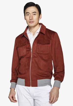 Mężczyzna w rdzawo-brązowej kurtce na suwak z szarymi mankietami i pasem w talii, noszący białą koszulę i białe spodnie, stojący na białym tle.