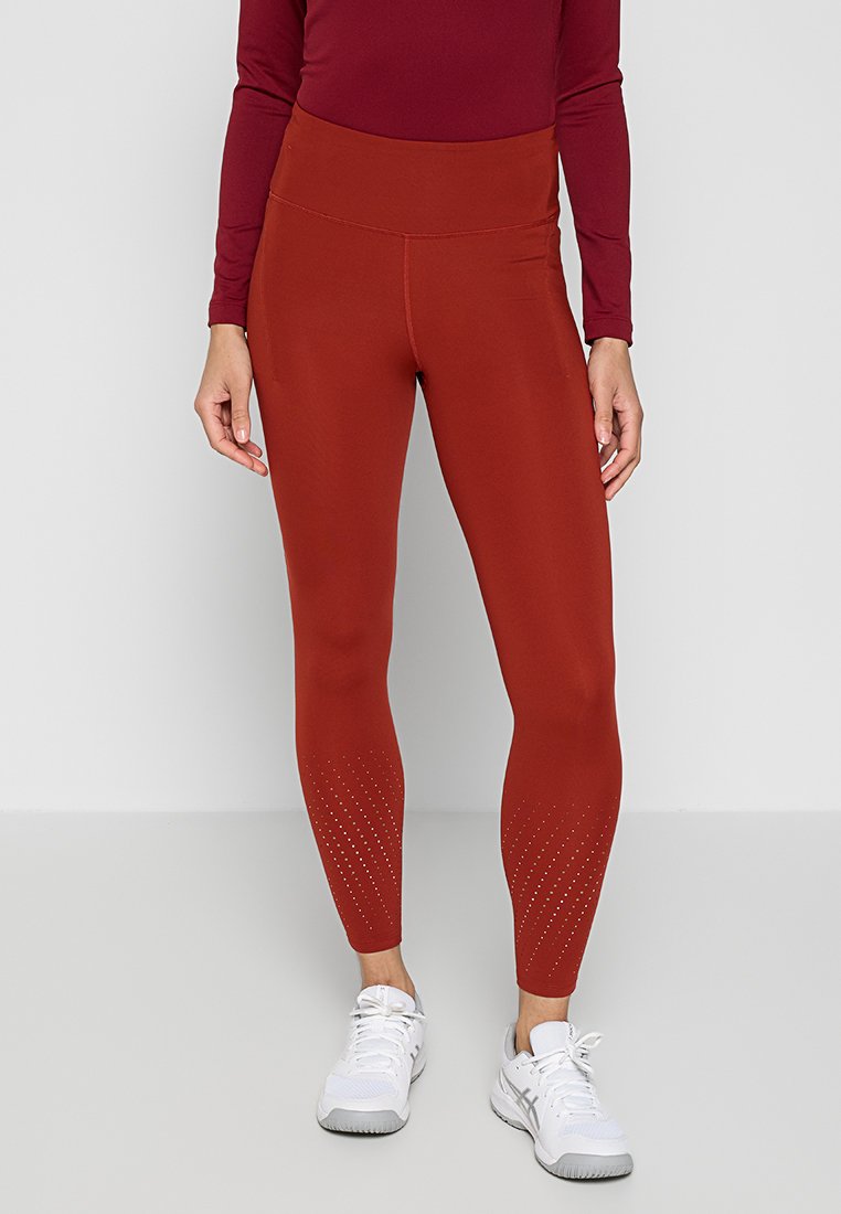 Leggings a vita alta di colore ruggine realizzati in tessuto elasticizzato, con dettagli perforati vicino alla caviglia. Abbinati a scarpe da ginnastica bianche.