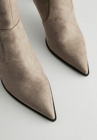 Botine bej din suede cu vârf ascuțit și talpă neagră. Partea superioară are cusături minime și o textură netedă.