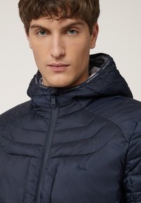 Navyblauer Puffer-Jacke mit Reißverschluss vorne, hohem Kragen und verstellbarem Kapuze. Mit Chevron-Steppnähten und einem kleinen gestickten Logo.