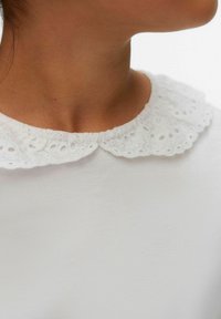 Top bianco in tessuto cotone a trama con un colletto in pizzo arrotondato. Il colletto presenta intricati motivi intagliati e un bordo arricciato.