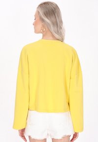 Žlutý zkrácený svetr s dlouhými rukávy, měkkou texturou a kulatým výstřihem, zkombinovaný s bílými roztrhanými šortkami.