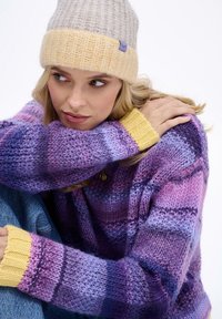 Fialovo-svetlofialový pruhovaný sveter s textúrovaným pleteným vzorom a žltými rebrovanými manžetami. V páre so sivou a žltou pletenou čiapkou.