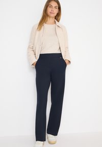 Navy blauwe stretchbroek met een aansluitende tailleband, gecombineerd met een beige textuurjasje en een lichte trui, aangevuld met neutrale sneakers.