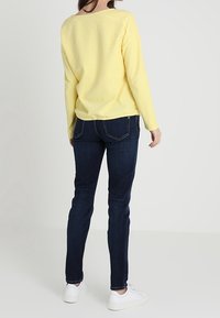 Gelber strukturierter Pullover, runder Ausschnitt, kombiniert mit dunkelblauen Slim-Fit-Jeans. Modell trägt weiße Sneakers, von hinten gesehen.