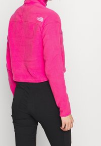 Różowa, krótka kurtka polarowa z wysokim kołnierzem i logo. Połączona z czarnymi, teksturowanymi spodniami z widocznymi kieszeniami.