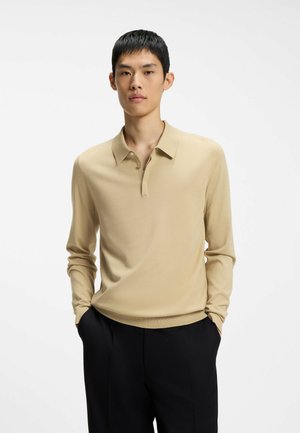 Ung mand med kort sort hår iført en beige langærmet polo og sorte bukser, stående med hænderne i lommerne mod en hvid baggrund.