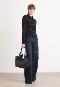 Zwarte lange mouwen shirt met knoopdetails, gecombineerd met wijde denim jeans. Model houdt een zwart leren handtas met gouden accenten vast.