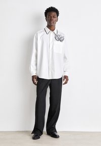 White button-up shirt with a hand-stitched floral design and black pocket; paired with loose black trousers and black shoes.