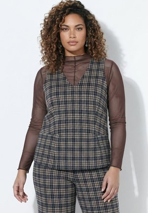 Femme aux cheveux bouclés portant des boucles d'oreilles argentées, un haut marron transparent à manches longues et un gilet à carreaux avec un pantalon assorti sur fond blanc.