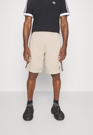 Mies pukeutuneena beigenvärisiin Adidas-shortseihin, joissa mustat sivuraitat, musta Adidas-paita valkoisilla raidallisilla hihoilla, mustat sukat ja mustat lenkkarit.