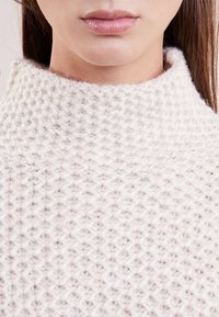 Pull en maille blanc cassé avec un col montant, présentant un motif texturé et un matériau doux et duveteux, idéal pour un porté décontracté et confortable.