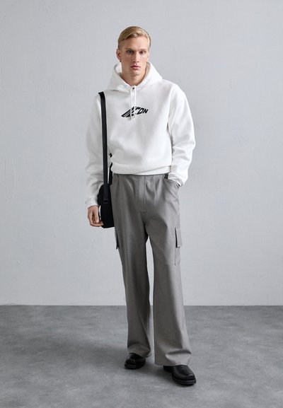 Sudadera blanca con logotipo negro, pantalones cargo grises de pierna ancha y zapatos negros. El modelo está de pie con una bolsa negra, destacando los detalles del atuendo.
