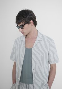 Camicia a maniche corte di un grigio chiaro con un motivo a zebra testurizzato, abbinata a una canotta verde spento. Occhiali da sole neri completano il look.