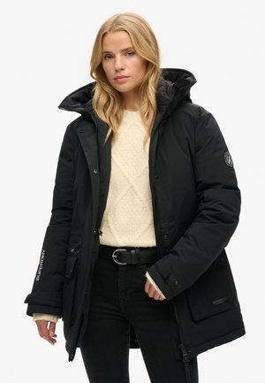 Abrigo acolchado negro con collar alto, cremallera y múltiples bolsillos. Llevado sobre un suéter crema y jeans negros, con detalles de marca en las mangas.