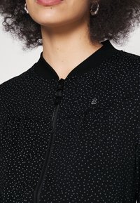 Femme portant un haut noir zippé avec petits pois blancs, col rond, et boucles d'oreilles créoles argentées.